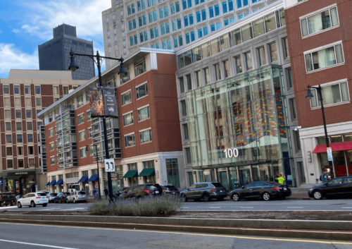Photograph of a city block showing the front of five story brick buildings surrounding a mostly glass and masonry center entrance. Taller buildings surround. People and cars lines the street and sidewalk in front.